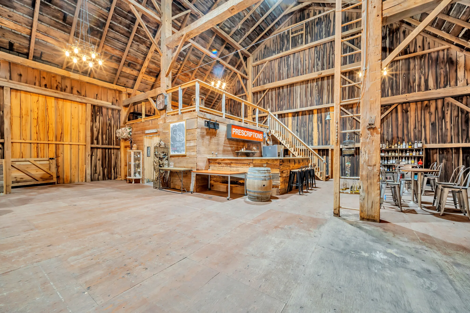 Inside the barn: weathered board walls and timber posts, a bar under a loft with a staircase, a sign reading PRESCRIPTIONS, high tables, a plank floor