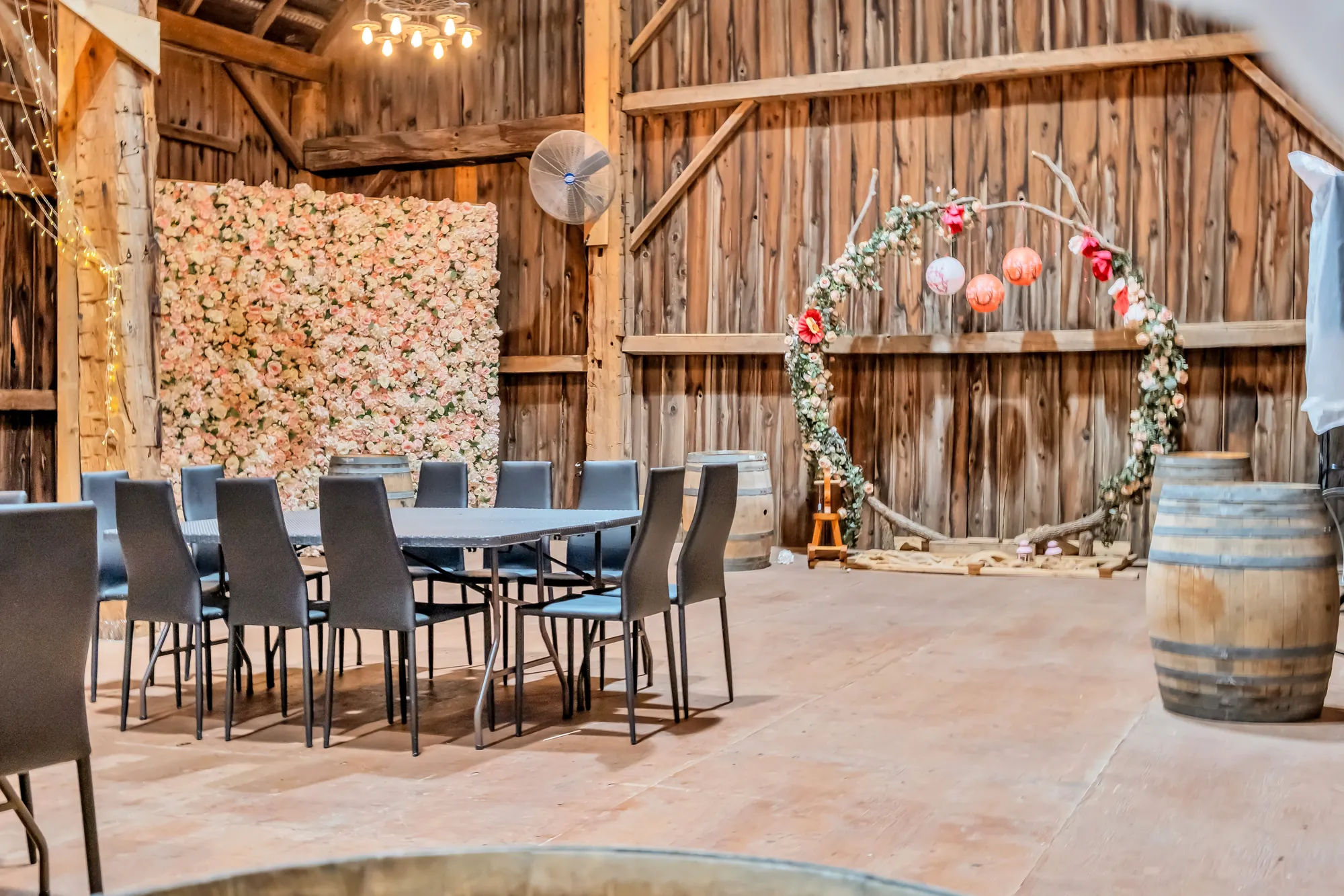 Two long tables with black chairs, a wall of flowers, a floral arch and wine barrels against the board walls
