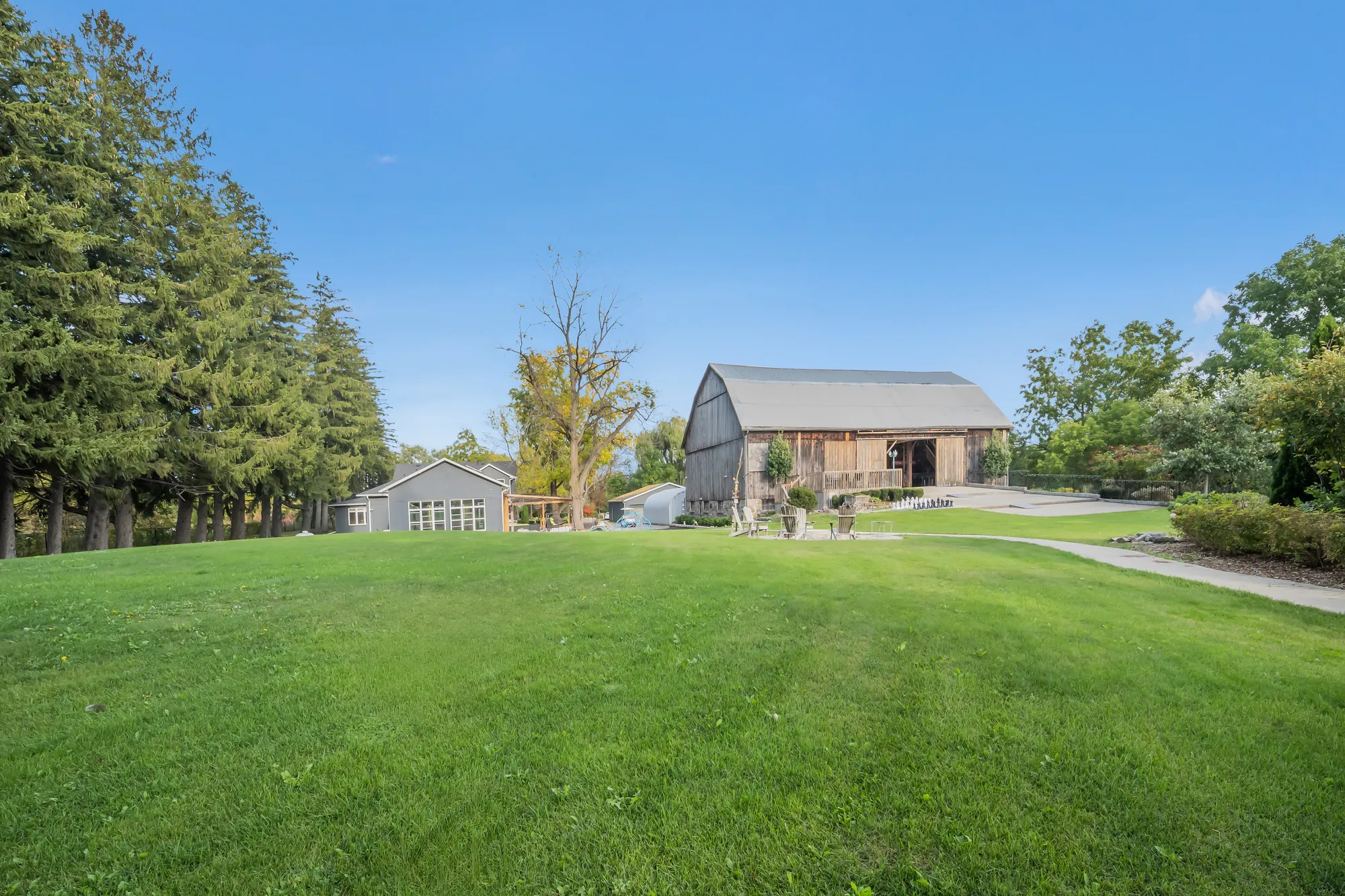 Across the lawn to a gambrel-roofed barn in weathered wood and a smaller grey building, spruce at the left