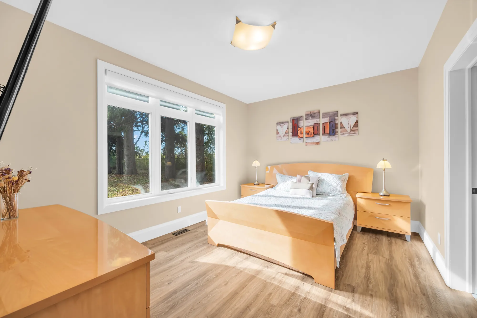 A bedroom with maple furniture: a bed with a white cover, night tables with lamps, a dresser, a window onto the trees