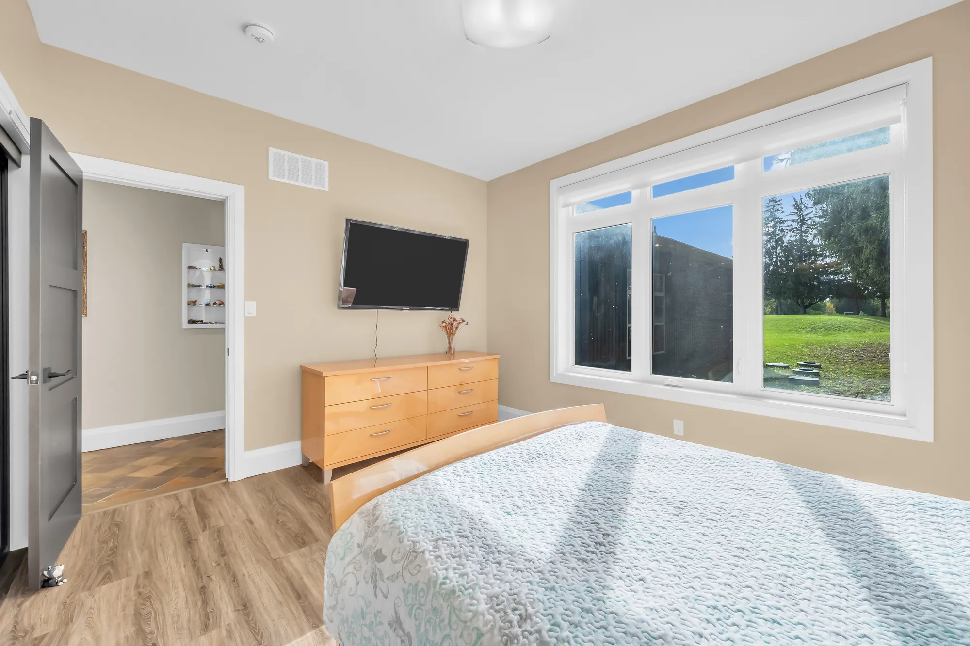 The same bedroom toward a wide window onto the lawn, a television on the wall, a maple dresser