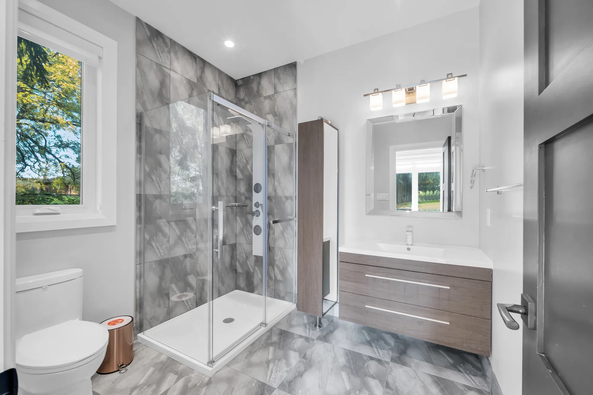 A bathroom with a glass-enclosed shower in grey tile, a floating vanity, a window