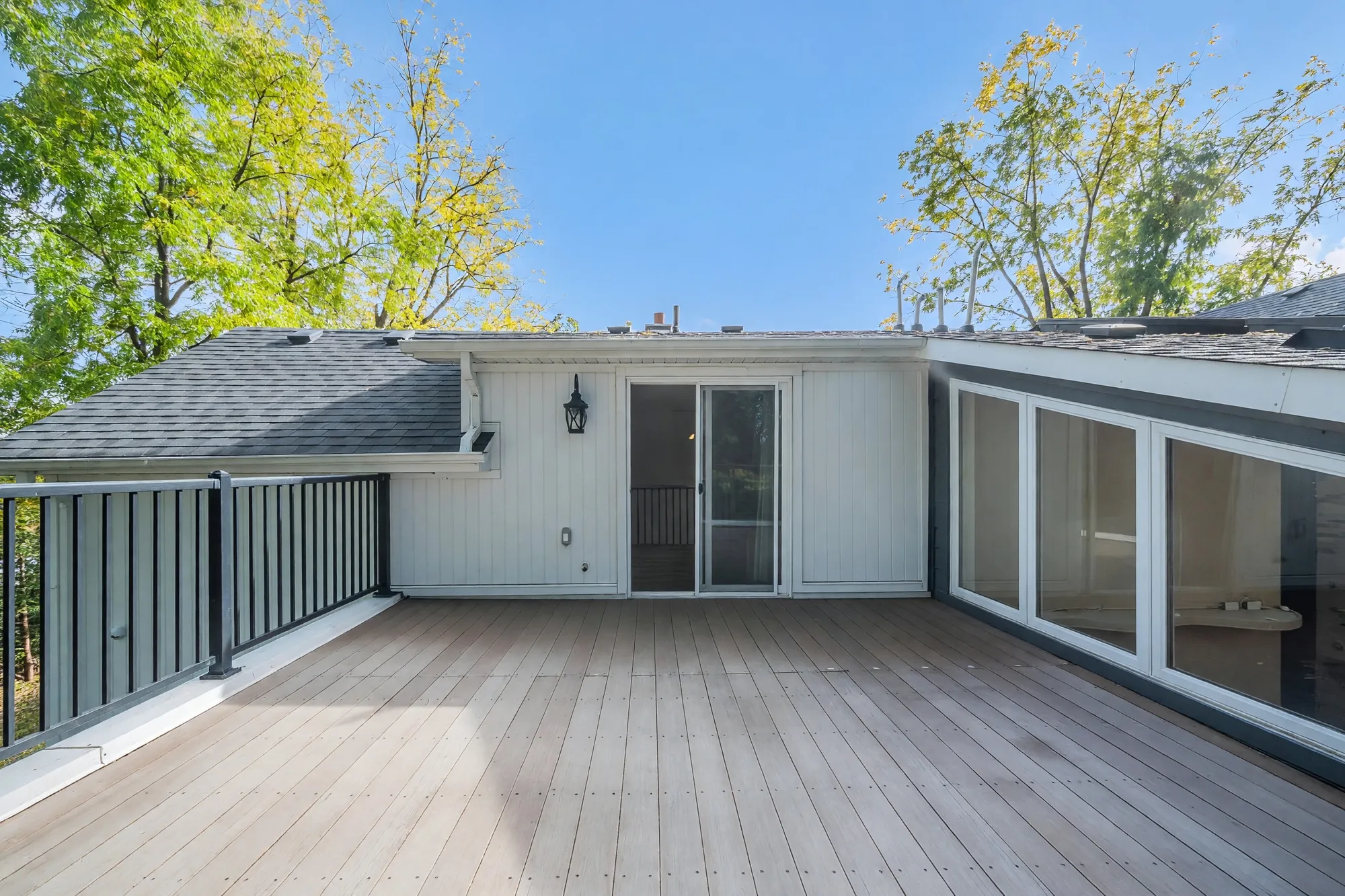 A balcony with weathered decking and a black railing, the white wall of the house with a sliding door and three large windows