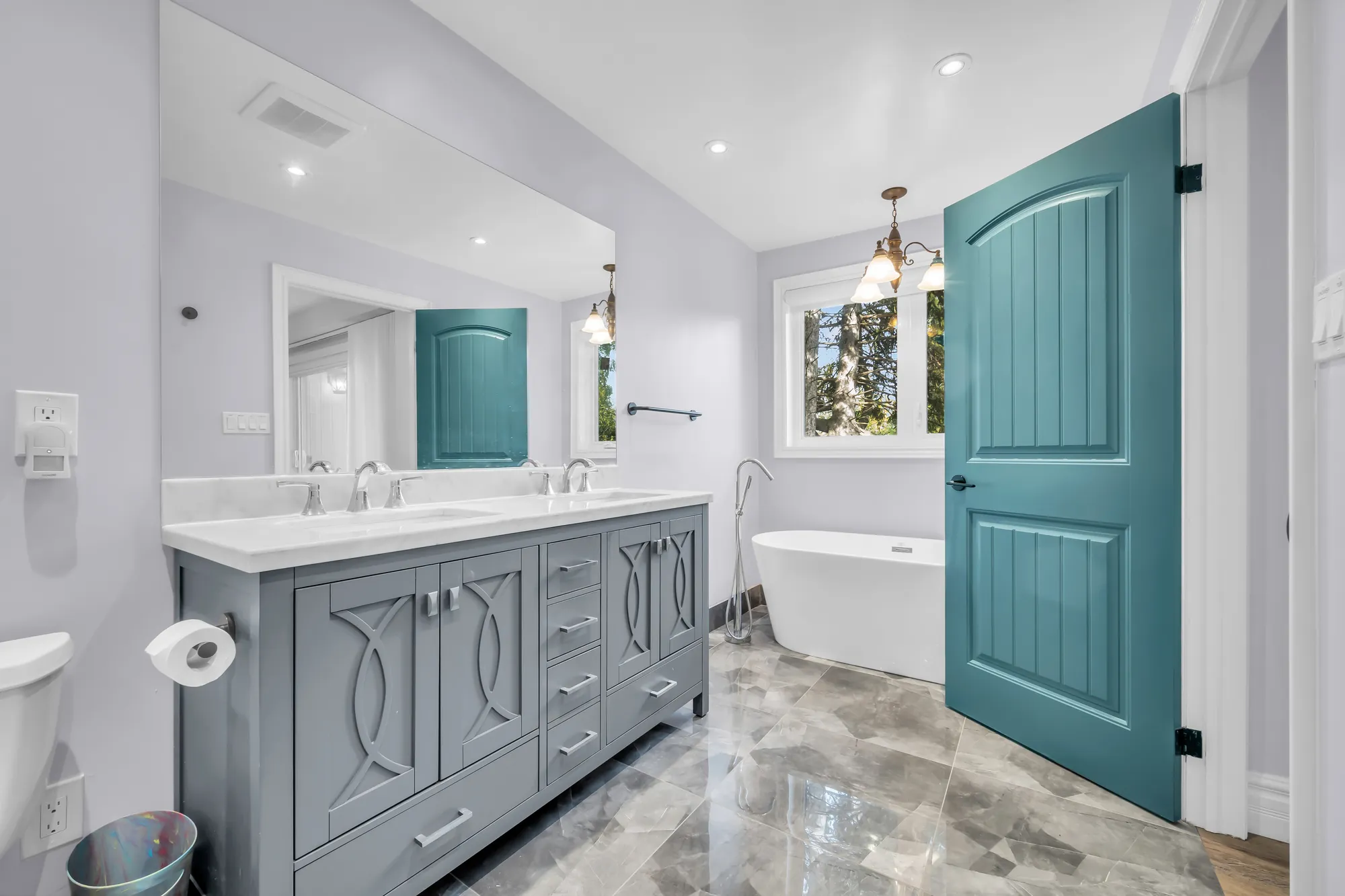 A bathroom: a double vanity in grey with ornamental doors, a freestanding tub under a window, marble-look tile, a teal door