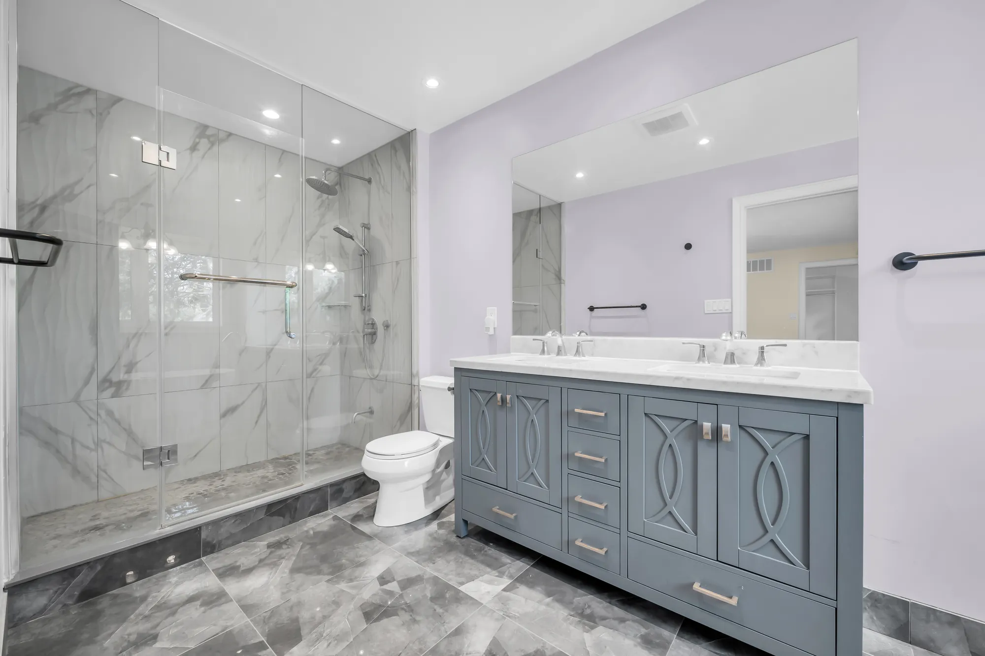 The same bathroom: a glass shower in marble-look tile, the double vanity, a toilet