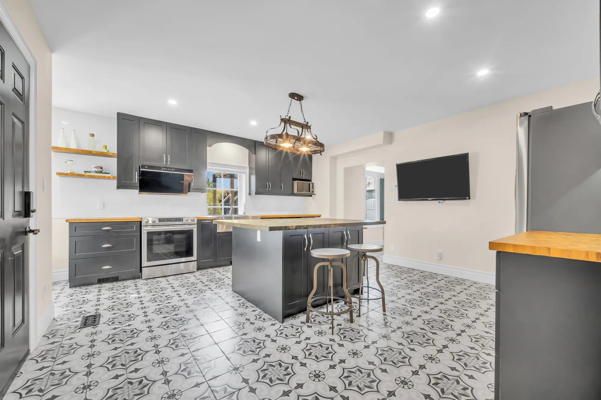 The same kitchen toward the range and a television on the wall, open shelves, stools at the island