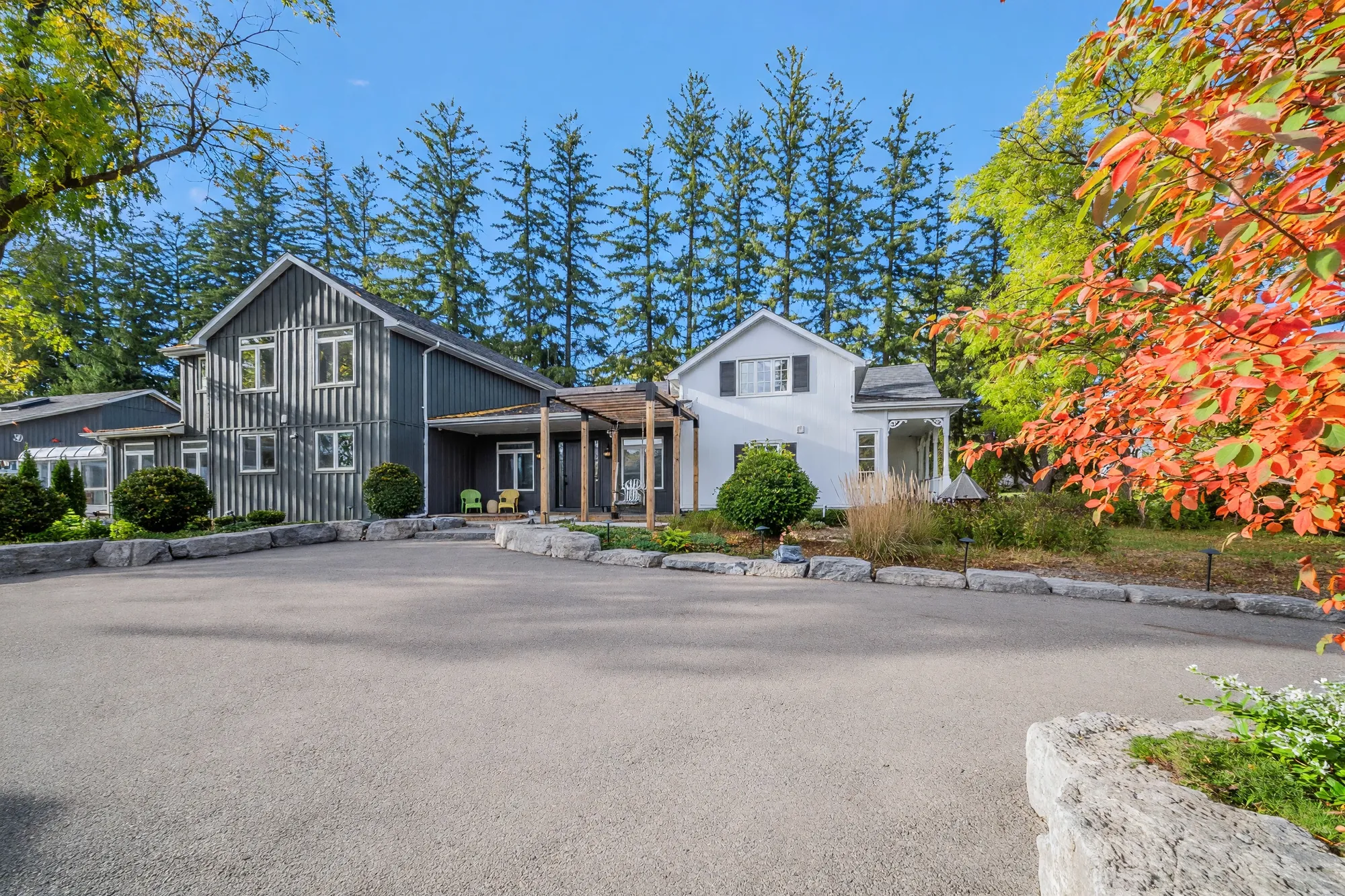 The front of the house across the paved forecourt: a two-storey wing in dark board-and-batten siding at the left, a porch under a cedar pergola in the middle, a white two-storey wing with dark shutters at the right, a row of tall spruce behind, a red maple at the right edge