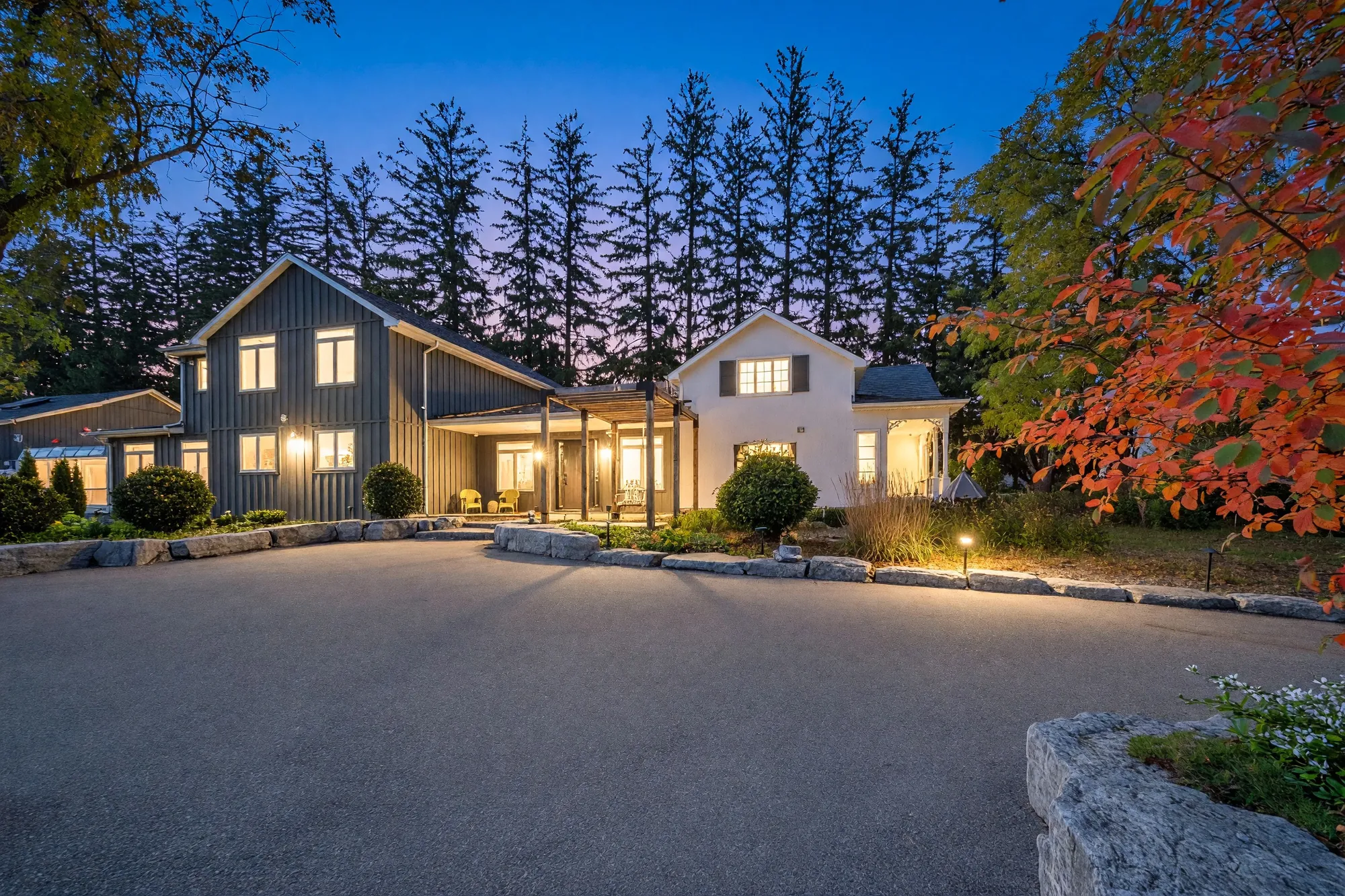 The front of 11584 Highway 48 at dusk: a two-storey wing in dark board-and-batten siding, a porch under a cedar pergola and a white two-storey wing, the windows lit, a row of tall spruce behind and a red maple at the right