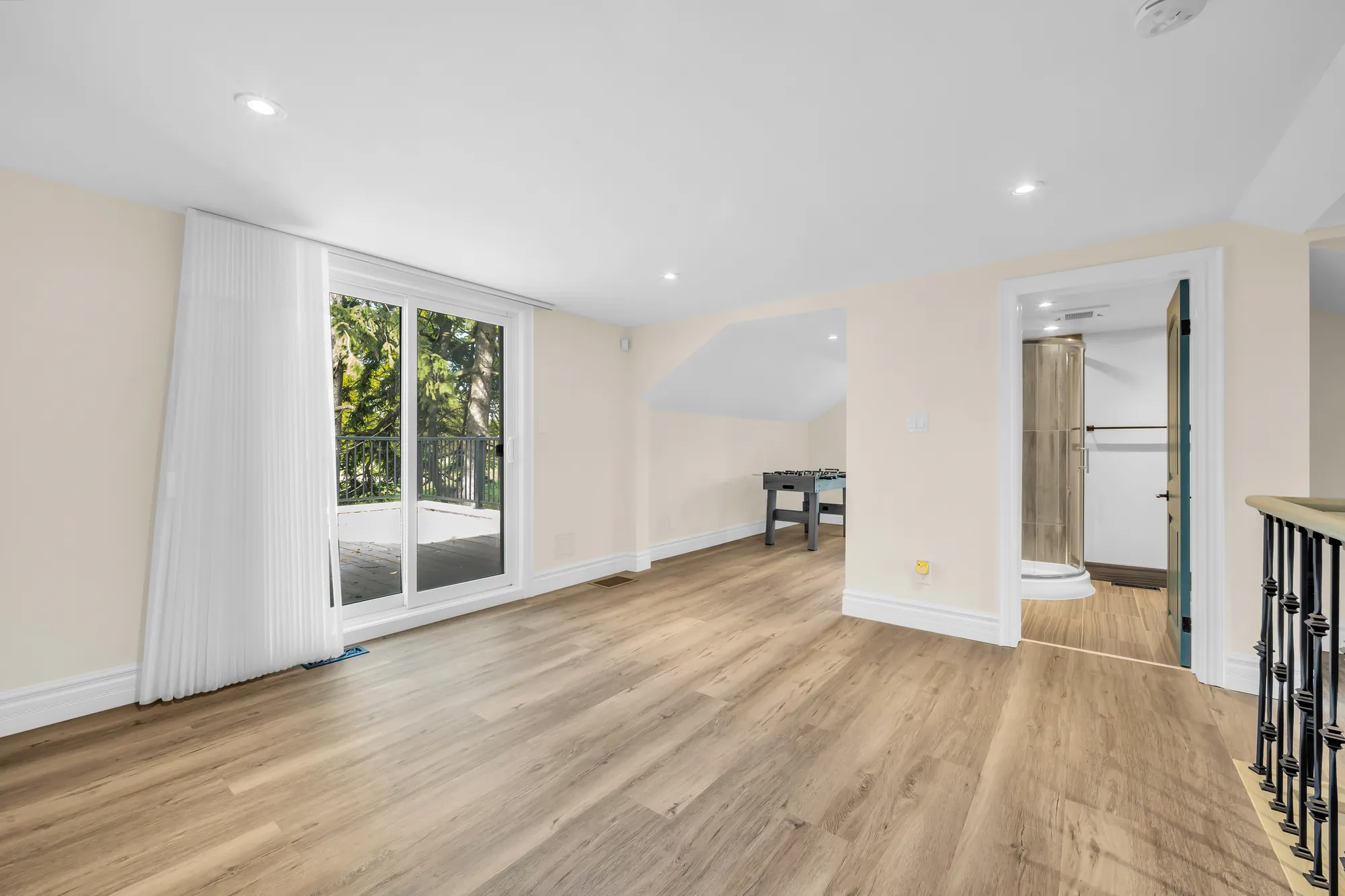 A long room upstairs in the east wing: sliding glass doors to a balcony, a stair railing, wood-look floors
