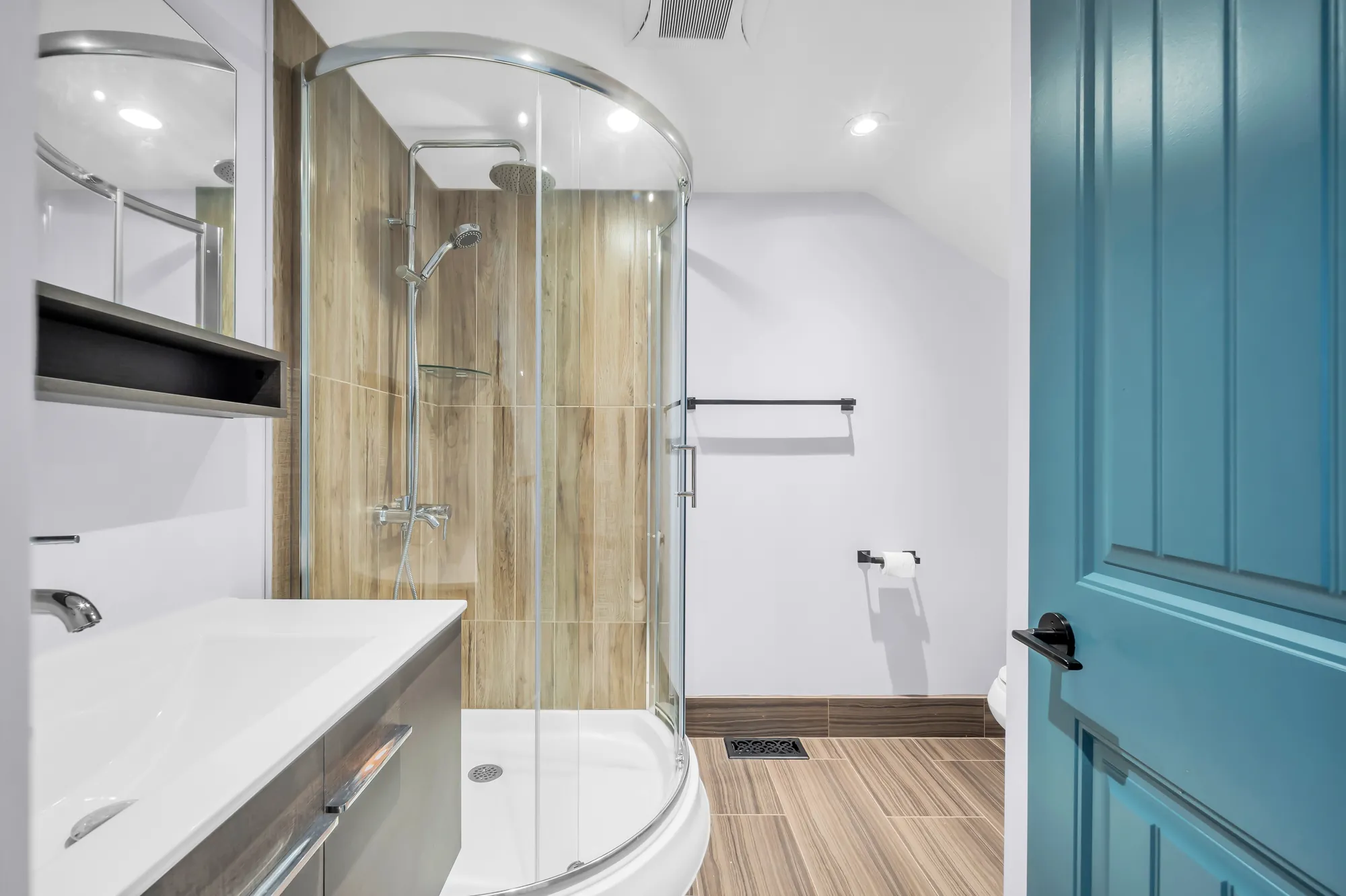 A bathroom: a curved glass shower in wood-look tile, a white vanity, a teal door