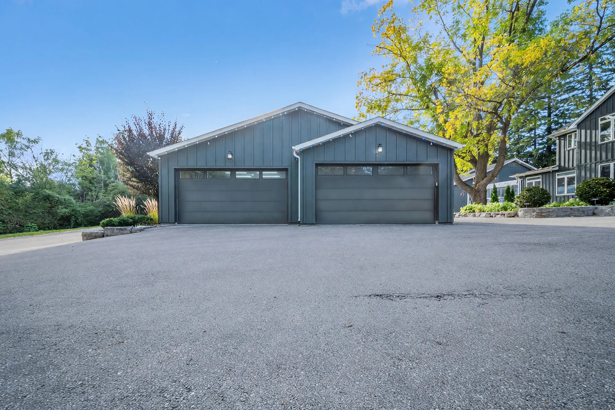 The detached garage: two gabled bays in dark board-and-batten siding with two wide garage doors, a paved apron, a large tree and the house at the right