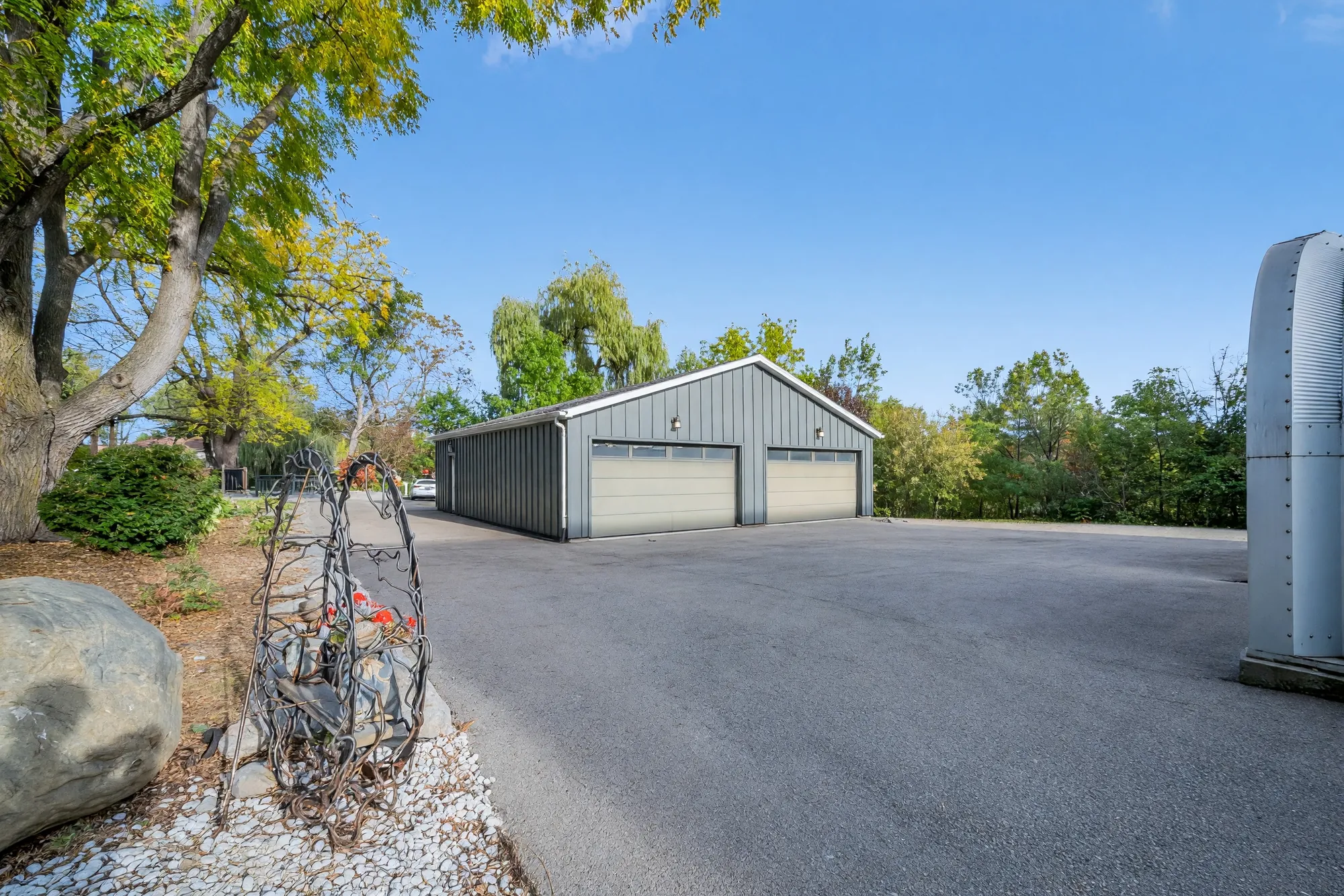 The drive toward the garage from beside the house: a boulder and a metal sculpture in a bed of white stone, trees around the garage