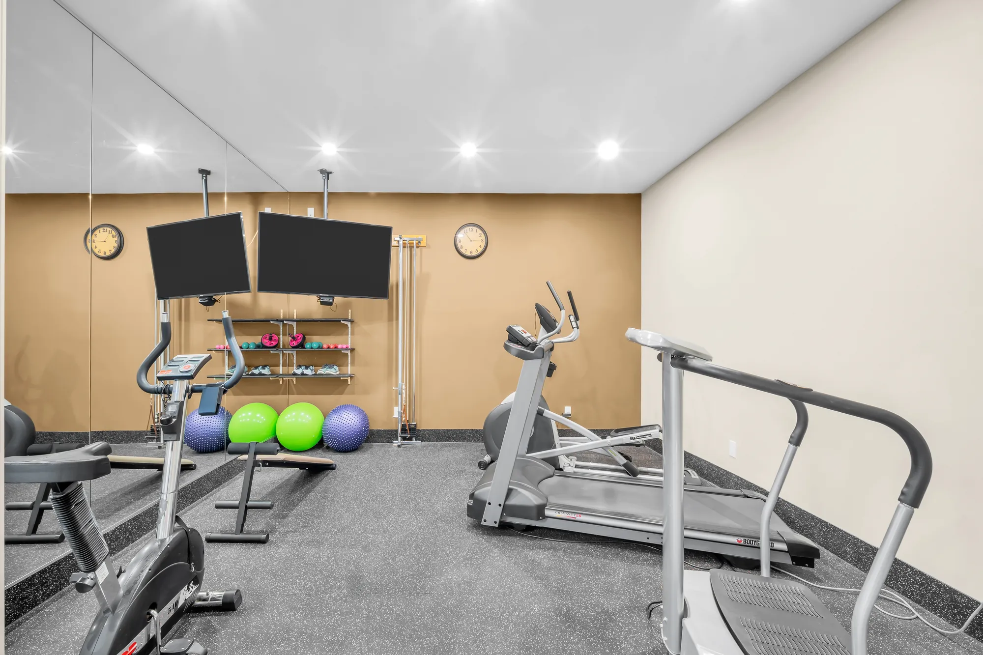 The gym: a treadmill, two exercise bikes, two televisions on the wall, a rubber floor, exercise balls