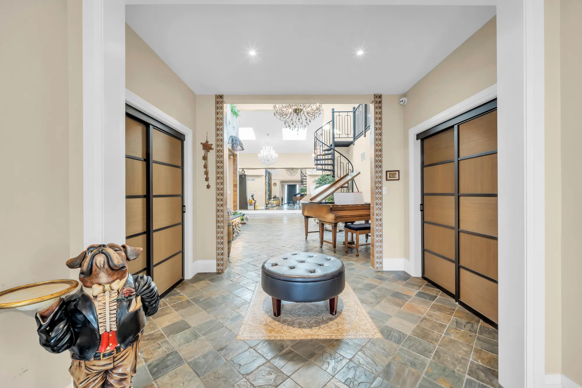 The hall from the front door: a slate floor, two closets with tall sliding doors, a round tufted ottoman, a carved bull statue, the flex area and a piano beyond