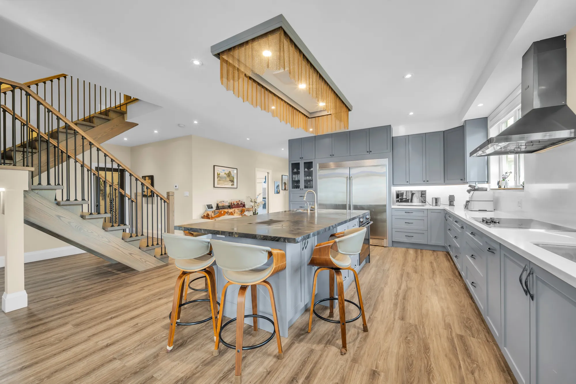 The kitchen: a large island in a dark veined stone with upholstered stools, grey shaker cabinets with glass-front uppers, stainless refrigerator columns, the wood stair at the left