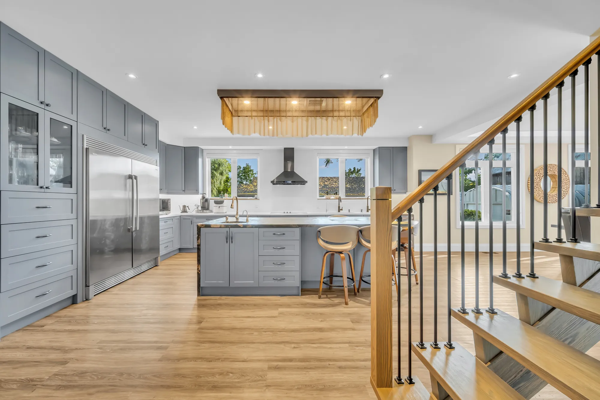 The kitchen toward the tall cabinets and the stainless refrigerator columns, a hood fan over the cooktop, the island and its stools, the stair at the right