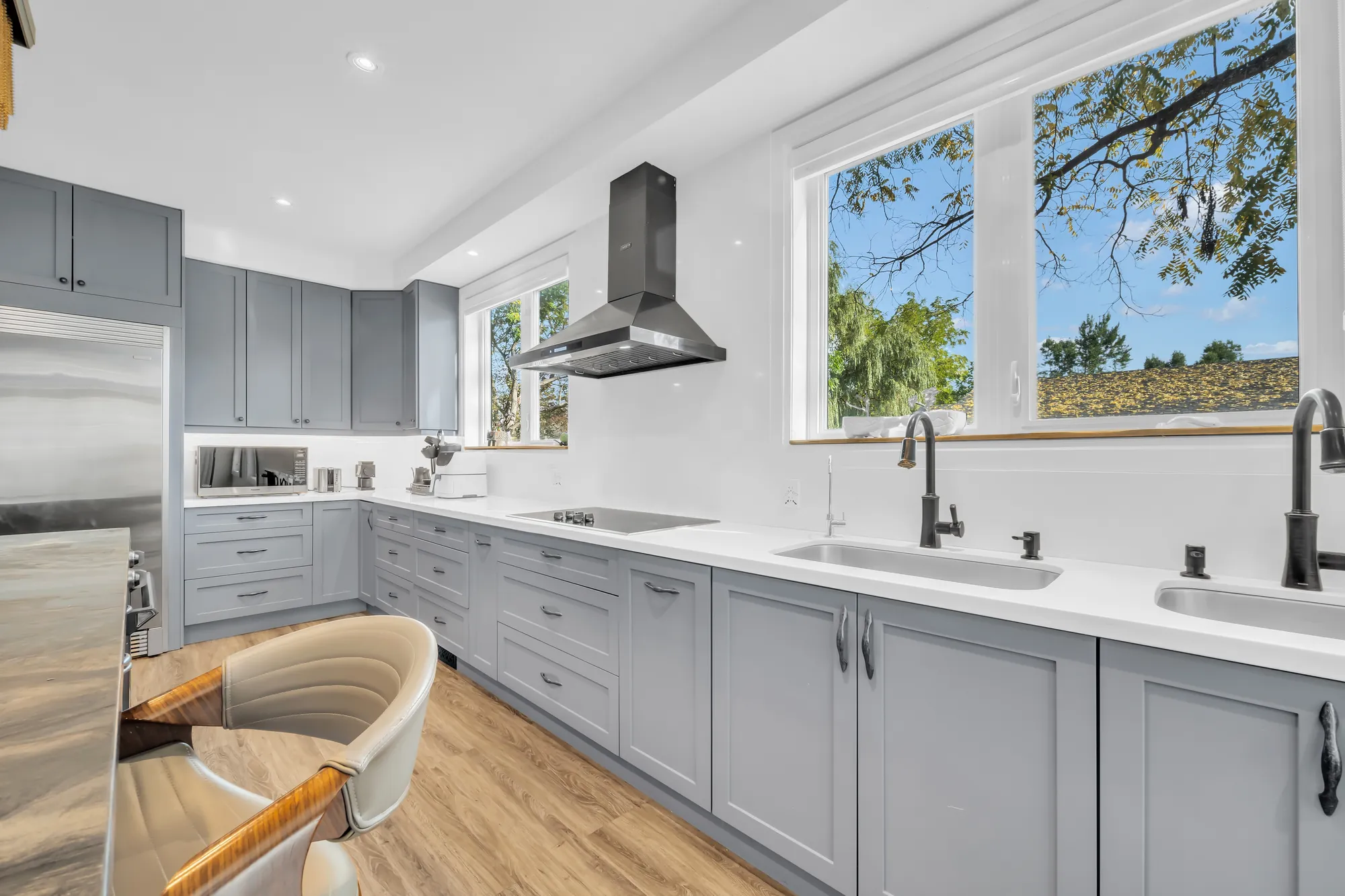 The counters under wide windows: two sinks, a cooktop under a stainless hood, grey shaker cabinets