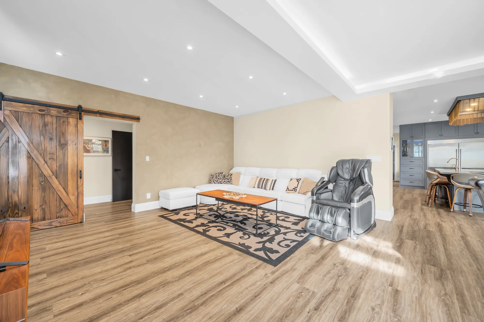 The living room: a white sectional and a coffee table on a patterned rug, a massage chair, a sliding barn door in old wood, the kitchen beyond