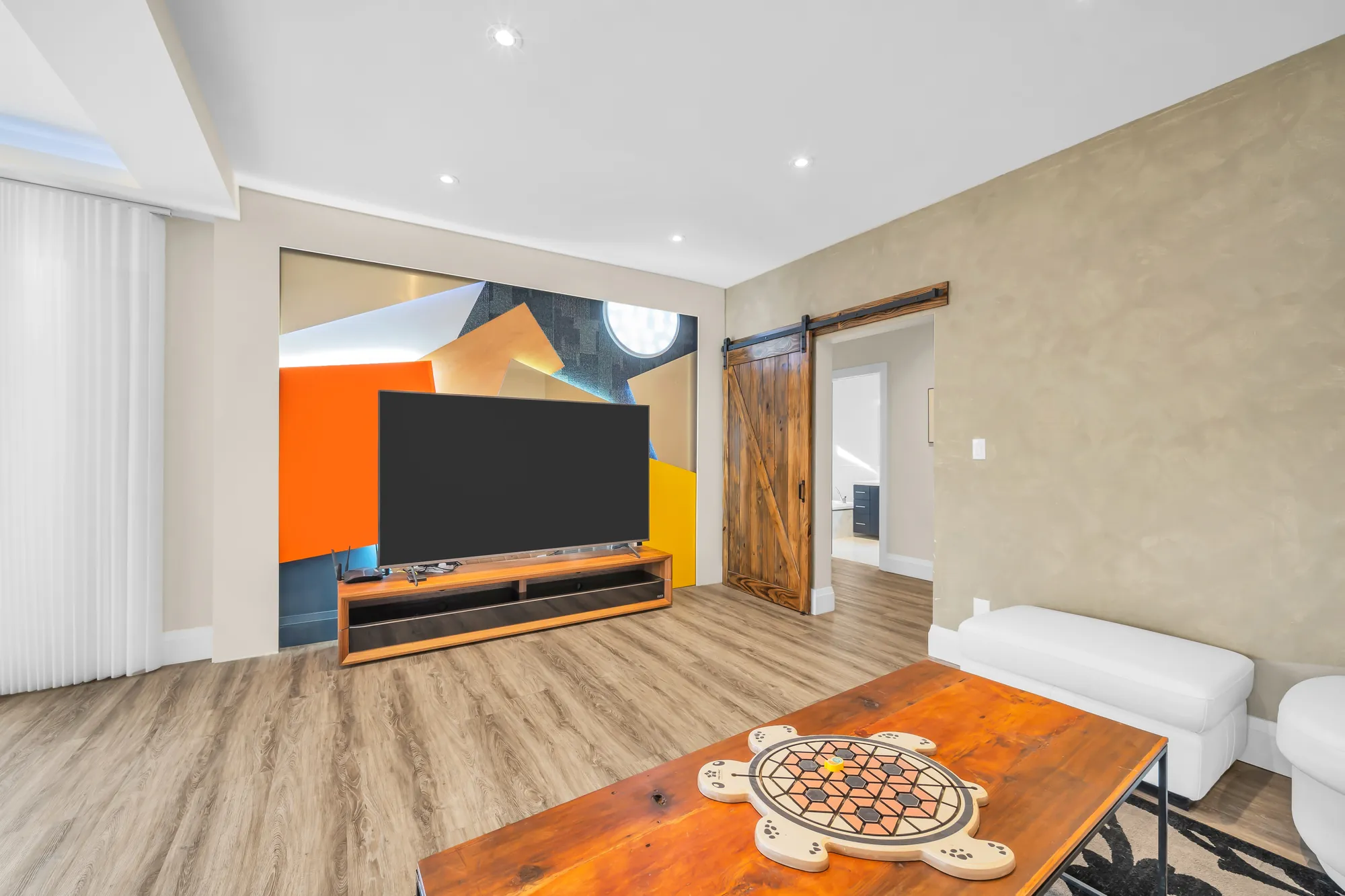 A television on an orange and grey mural wall, a sliding barn door, a carved wood table in the foreground