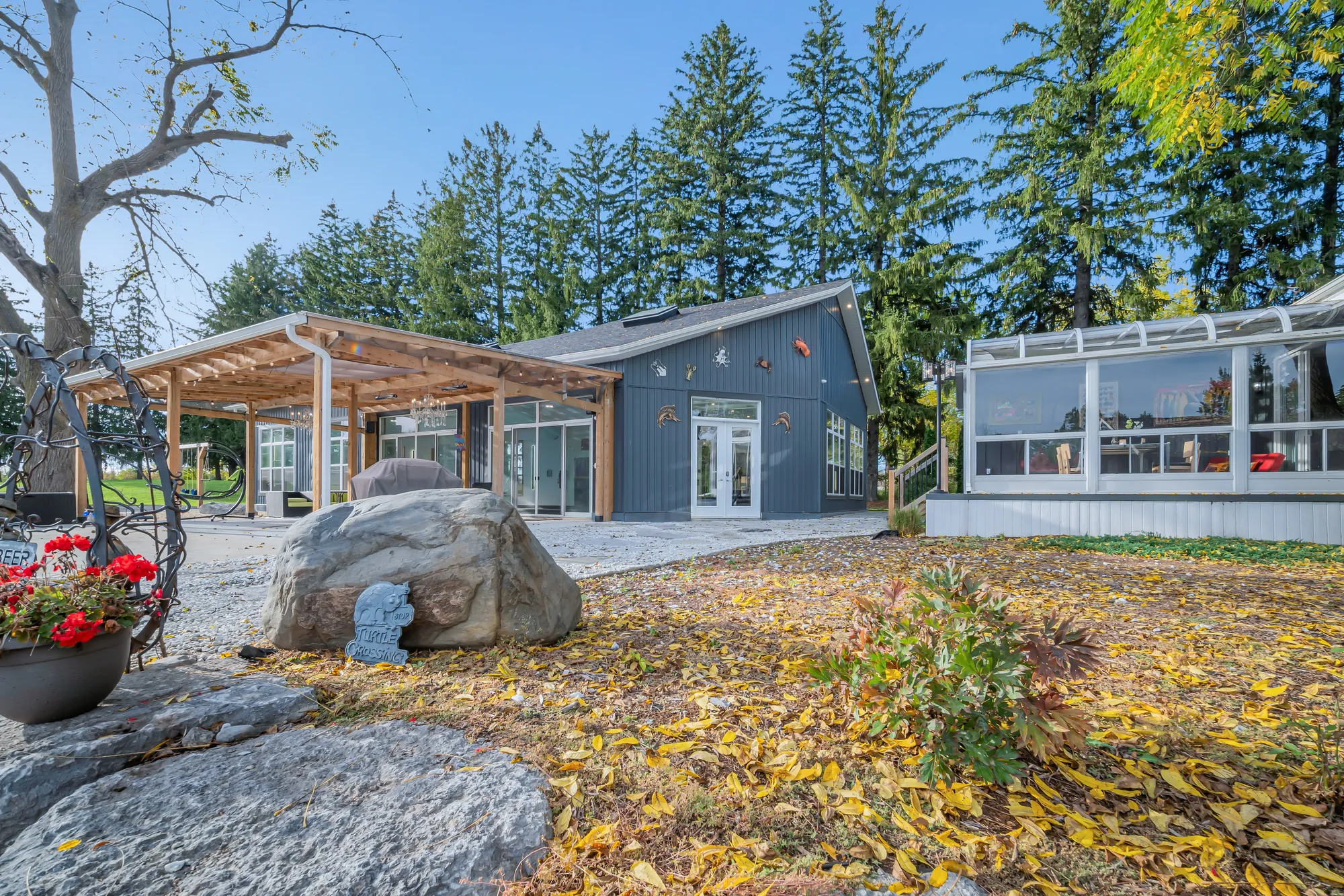 The patio side: a cedar pergola, the single-storey part in dark siding with French doors, the glass sunroom at the right, a boulder and red geraniums in the foreground