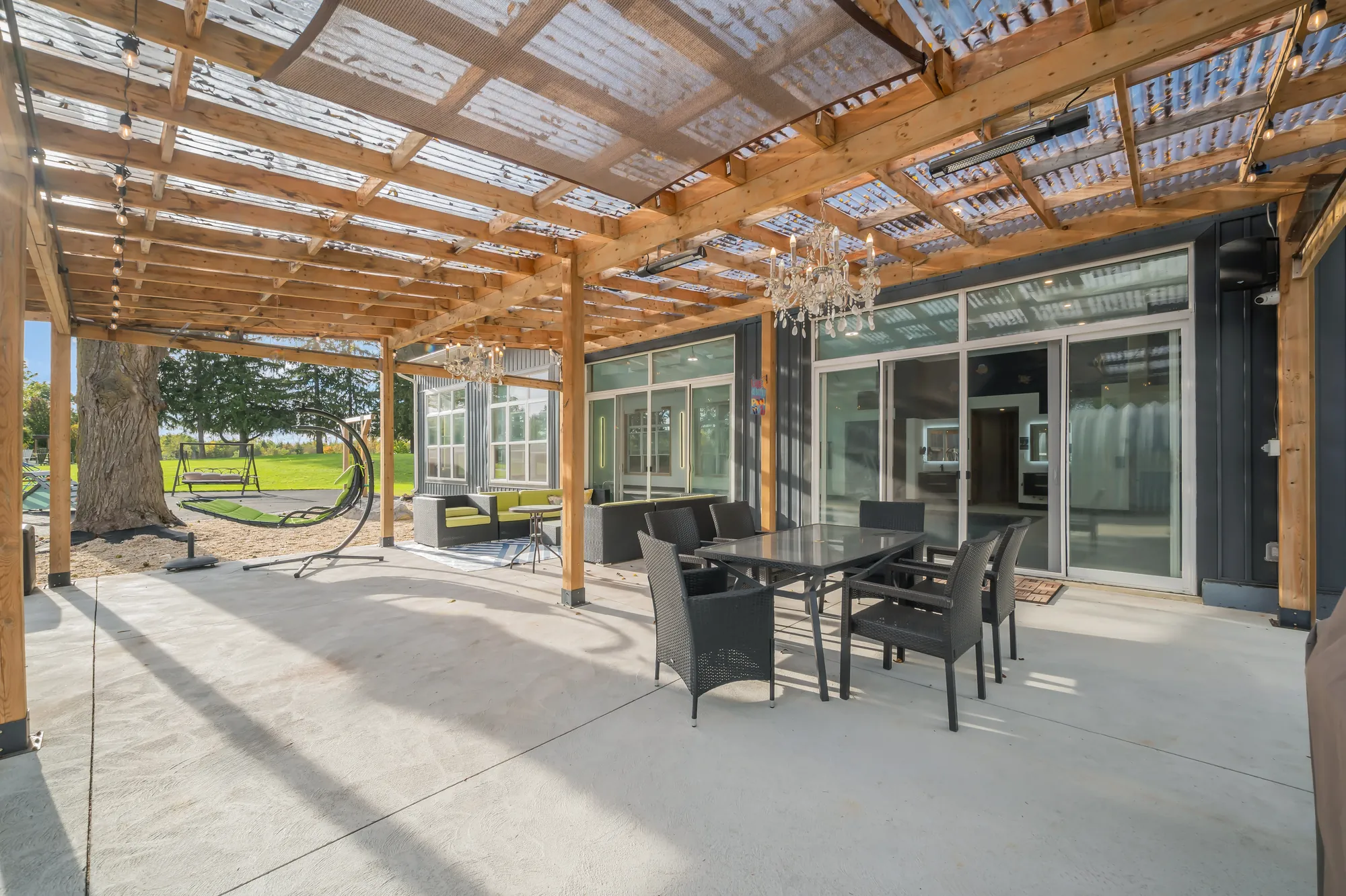 Under the pergola: a clear roof on cedar beams, a crystal chandelier, an outdoor dining table and chairs, a sectional sofa, a hanging chair, sliding glass doors into the house