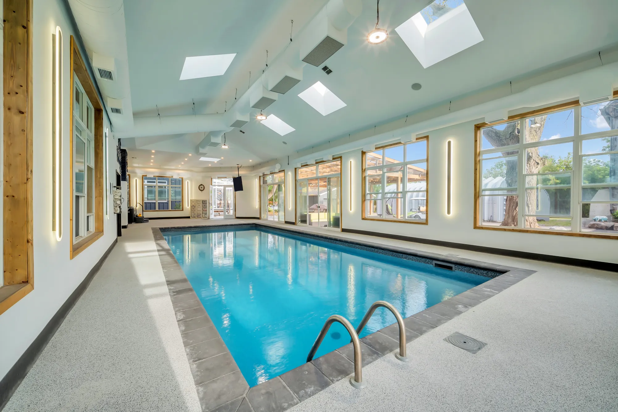 The indoor pool lengthwise: a long rectangular pool with a ladder, skylights, wood-framed windows, a speckled floor