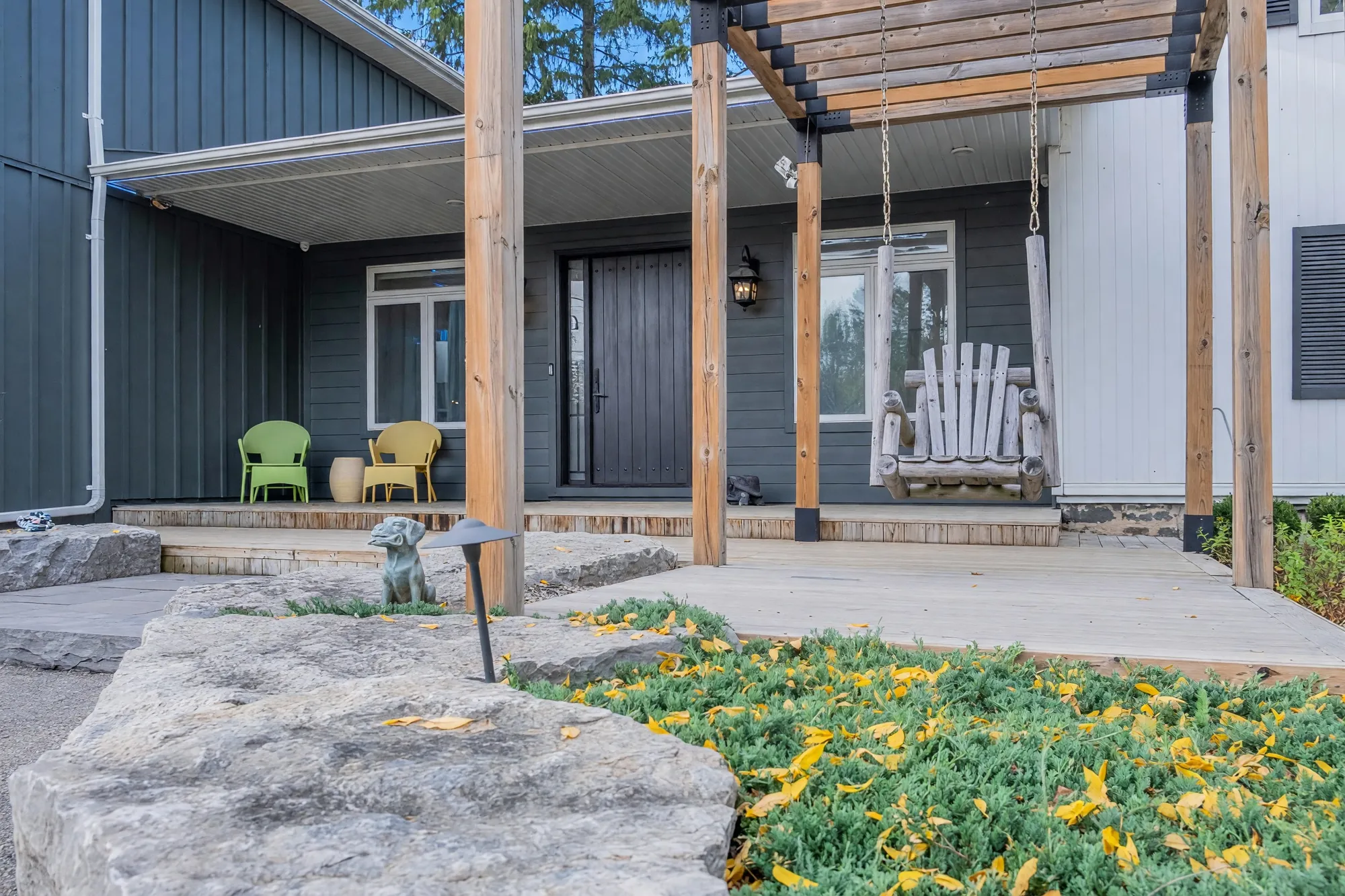The porch under the cedar pergola: a wooden porch swing on chains, two wicker chairs, the front door, limestone steps, a dog statue in the planting