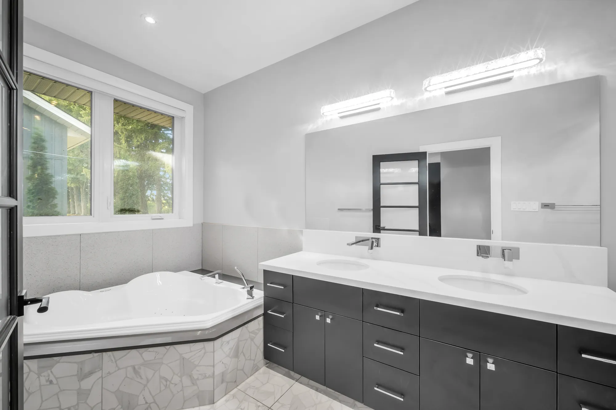The primary bathroom: a corner tub in grey tile, a long double vanity with dark cabinets, a wide mirror and lights
