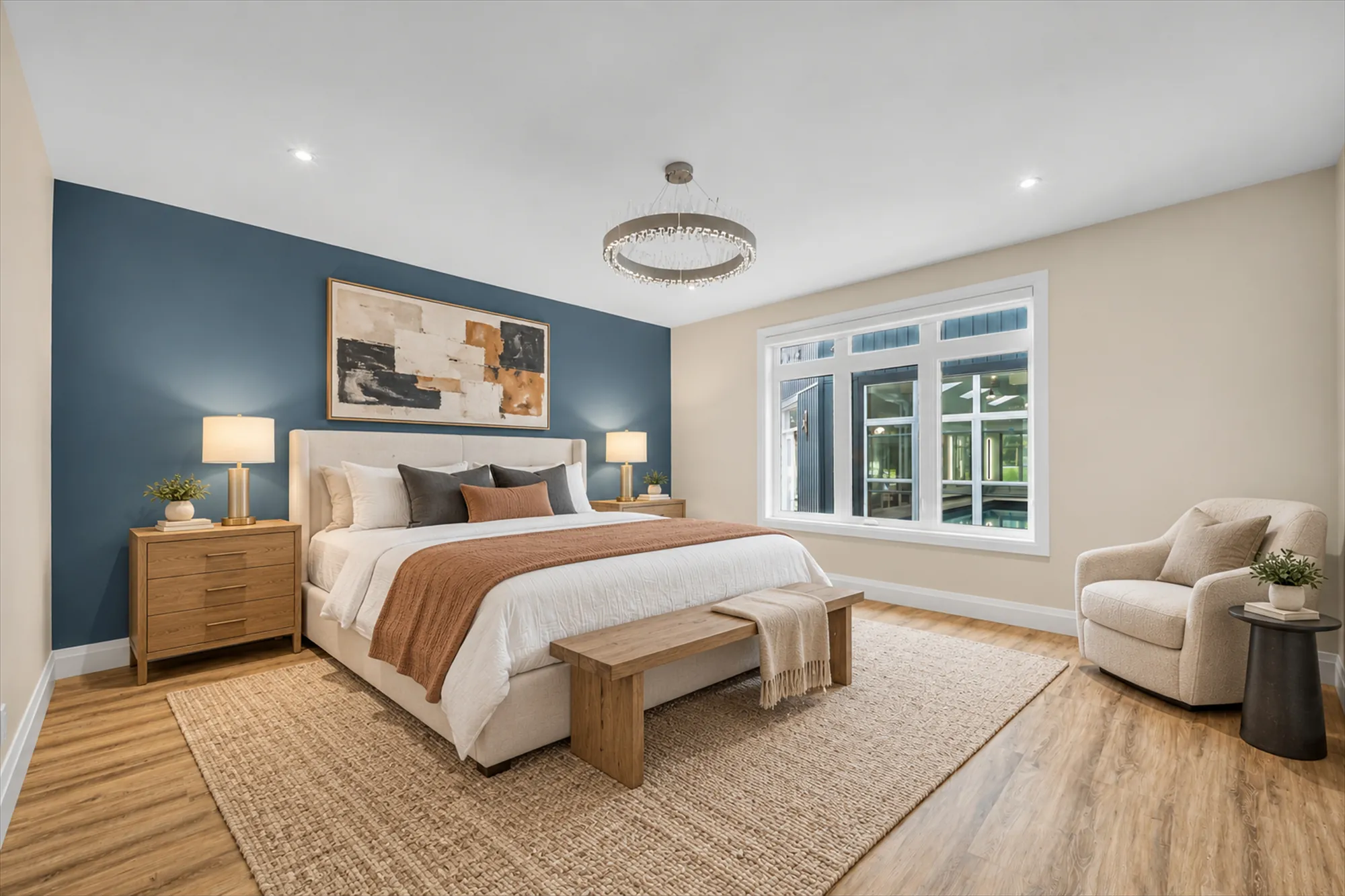 A bedroom with a dark blue accent wall, virtually staged: an upholstered bed and a bench, oak night tables and lamps, a jute rug, an armchair, a large abstract print, a crystal ring chandelier, a wide window