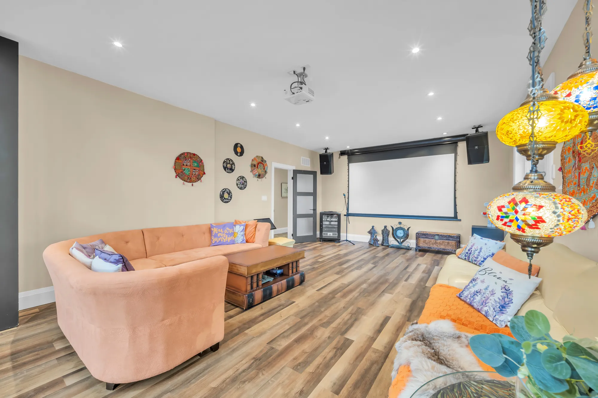 The screen end of the recreation room: a projector screen, orange sectional sofas, a trunk for a table, coloured glass lanterns
