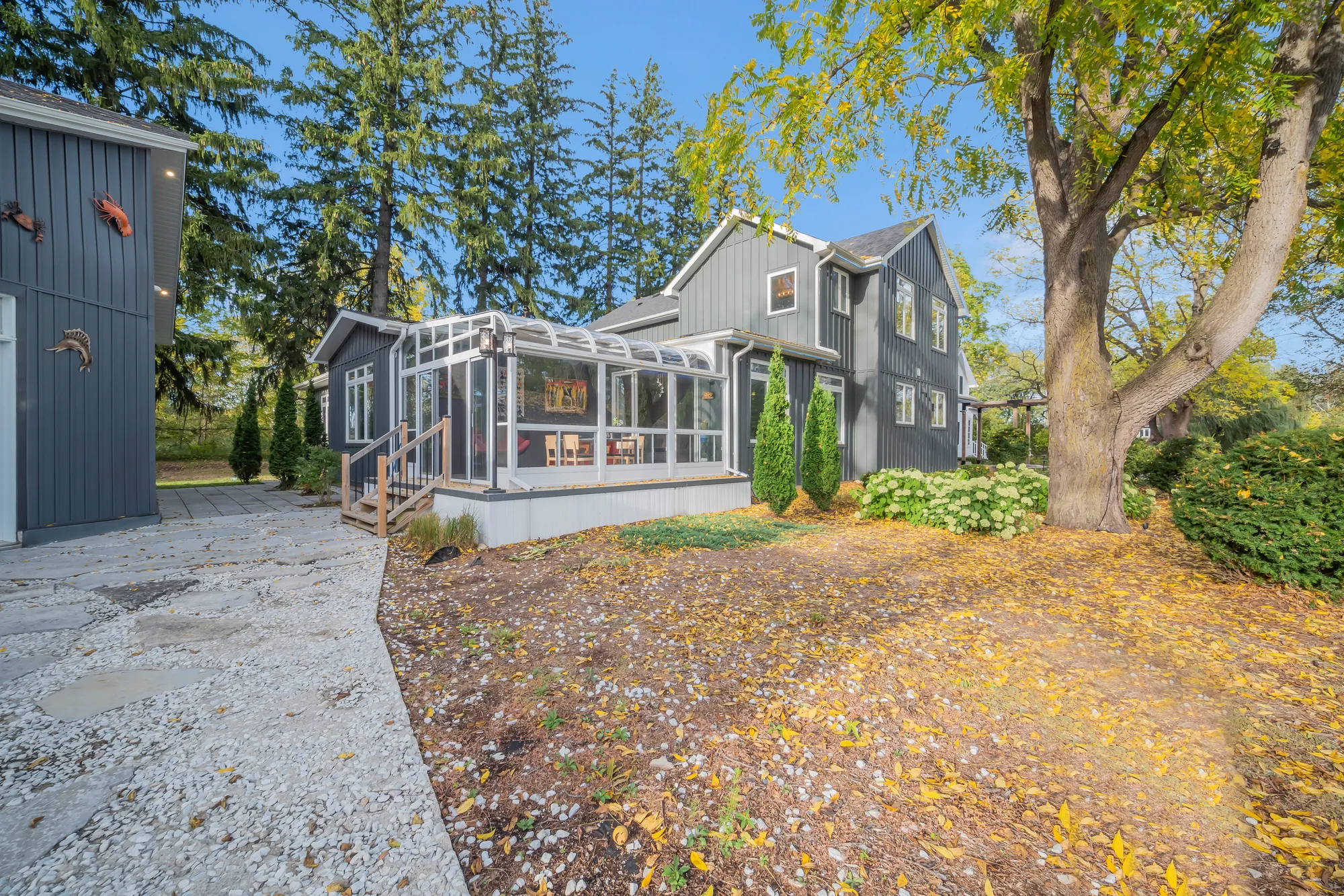 The side of the house: a glass sunroom on a deck, the dark two-storey wing behind it, a walk of crushed stone, fallen leaves on the garden bed