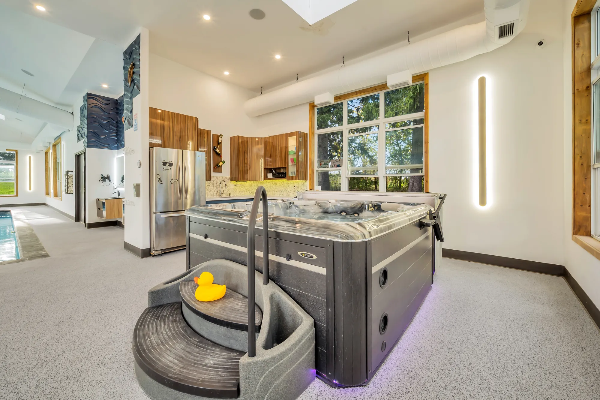 A hot tub with steps at one end of the pool room, a kitchenette with wood cabinets and a refrigerator beyond