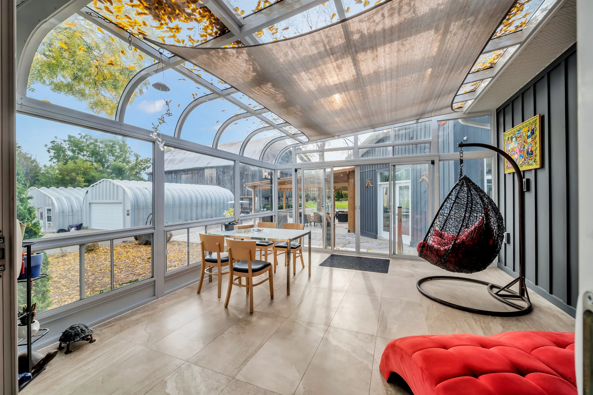 The sunroom: a curved glass roof on a white frame, glass walls, a dining table and chairs, a hanging egg chair, a red chaise, the dark siding of the house wall