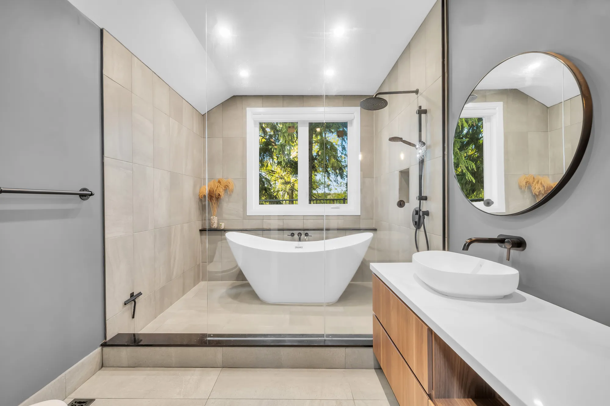 A bathroom: a freestanding tub under a window, a round mirror, a vessel sink on a wood vanity, grey tile