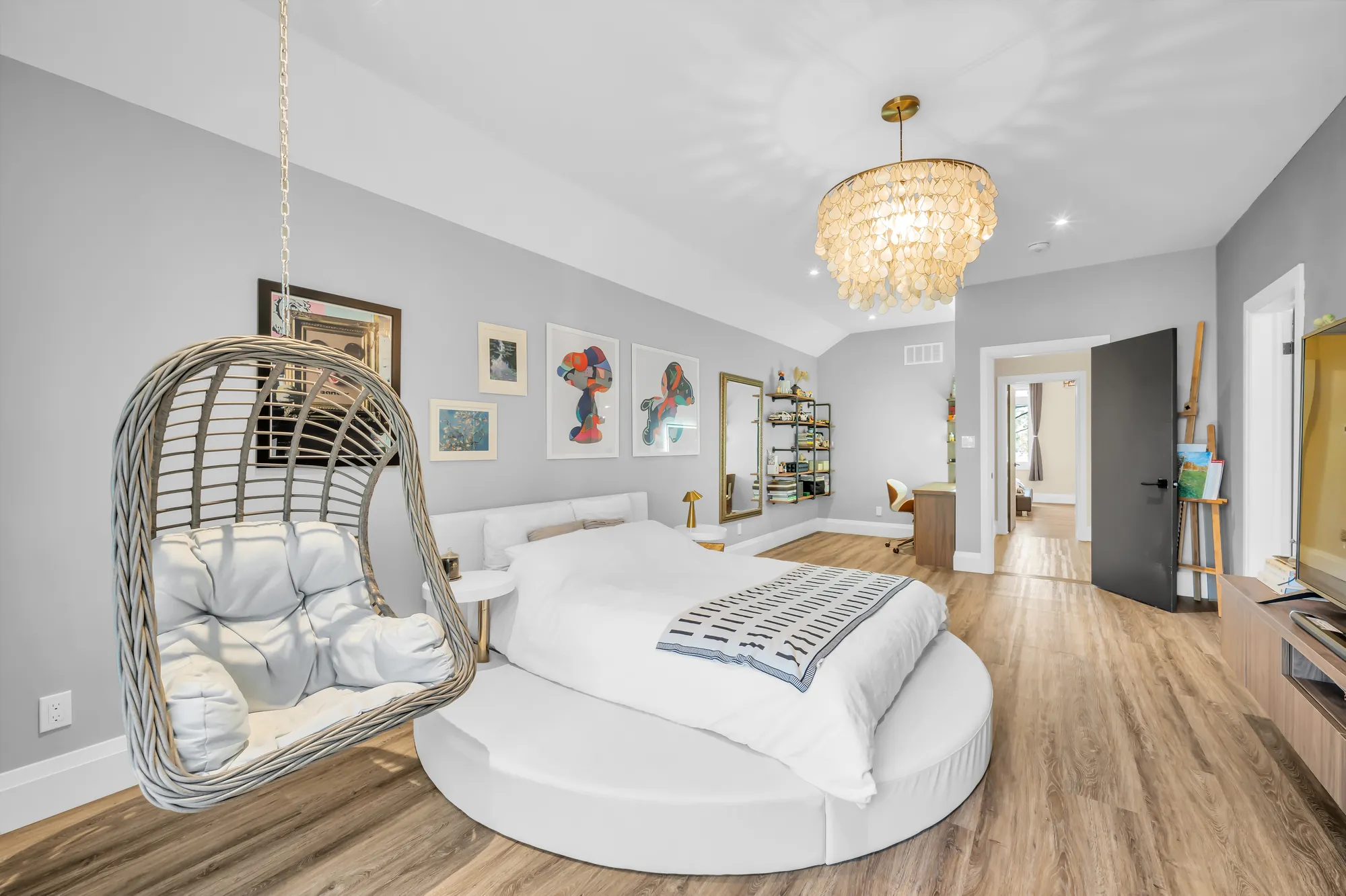 A bedroom with a round bed, a hanging rattan chair, framed prints, a crystal chandelier, a doorway to the hall