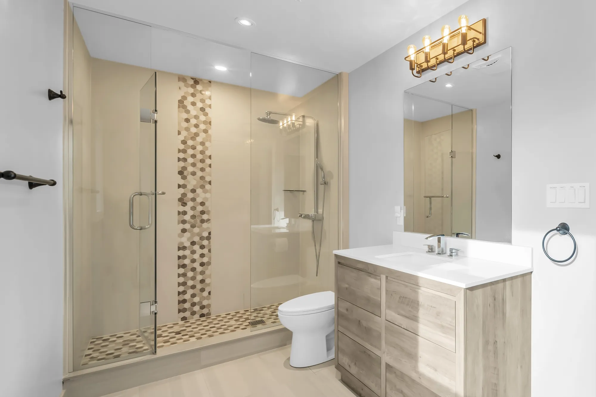 A bathroom: a glass shower with a strip of hexagon mosaic, a toilet, a wood-look vanity, brass lights
