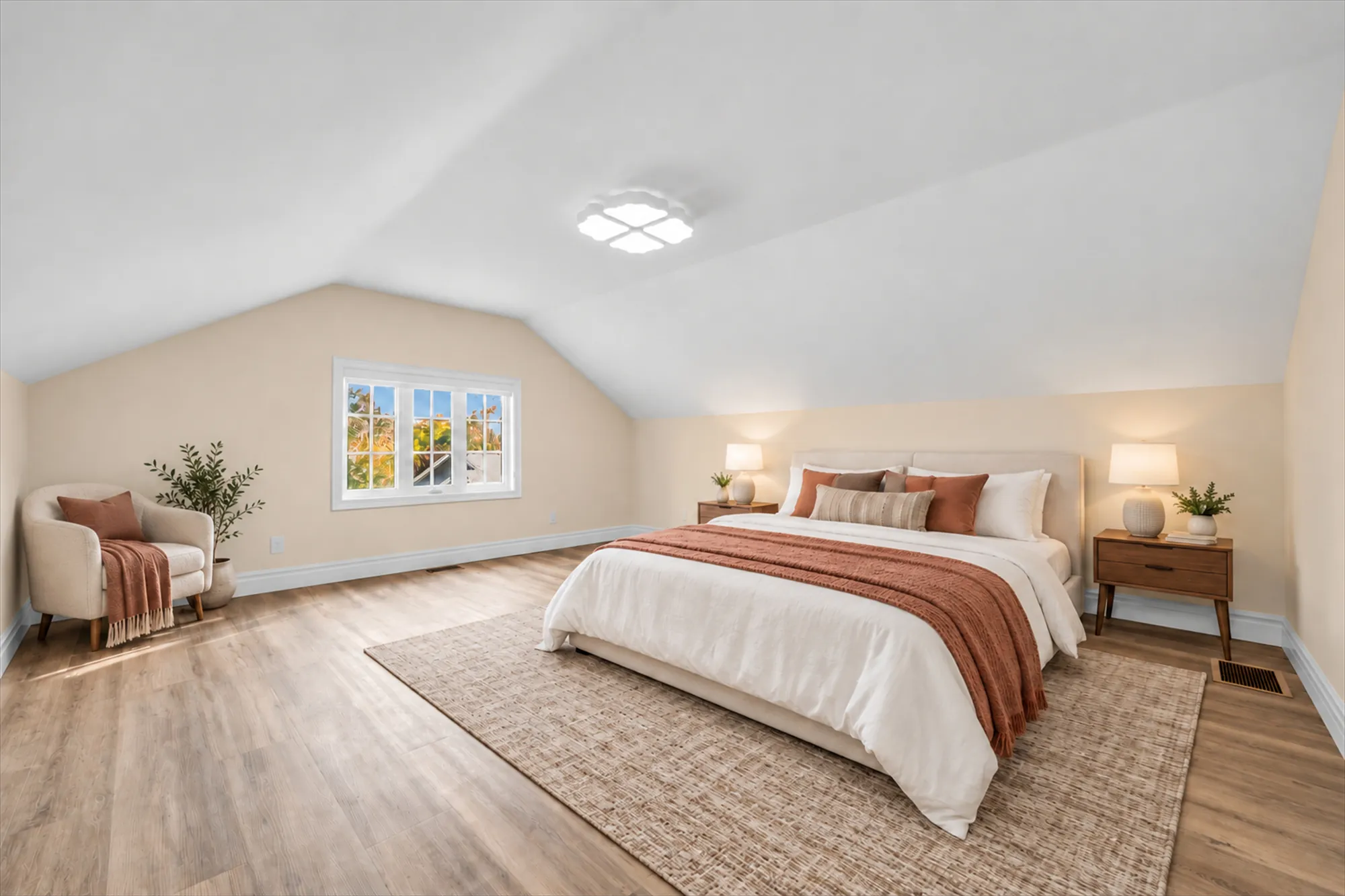 A bedroom under a sloped ceiling with a window in the gable, virtually staged: a bed with a rust throw, night tables and lamps, an armchair, a rug and a plant